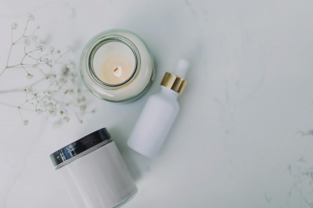 Overhead view of spa essentials including a candle, dropper bottle, and jar on a white marble surface.